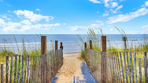 Ein Strandweg mit Holzzaun und blauer Bodenbelag, der zum Meer führt.