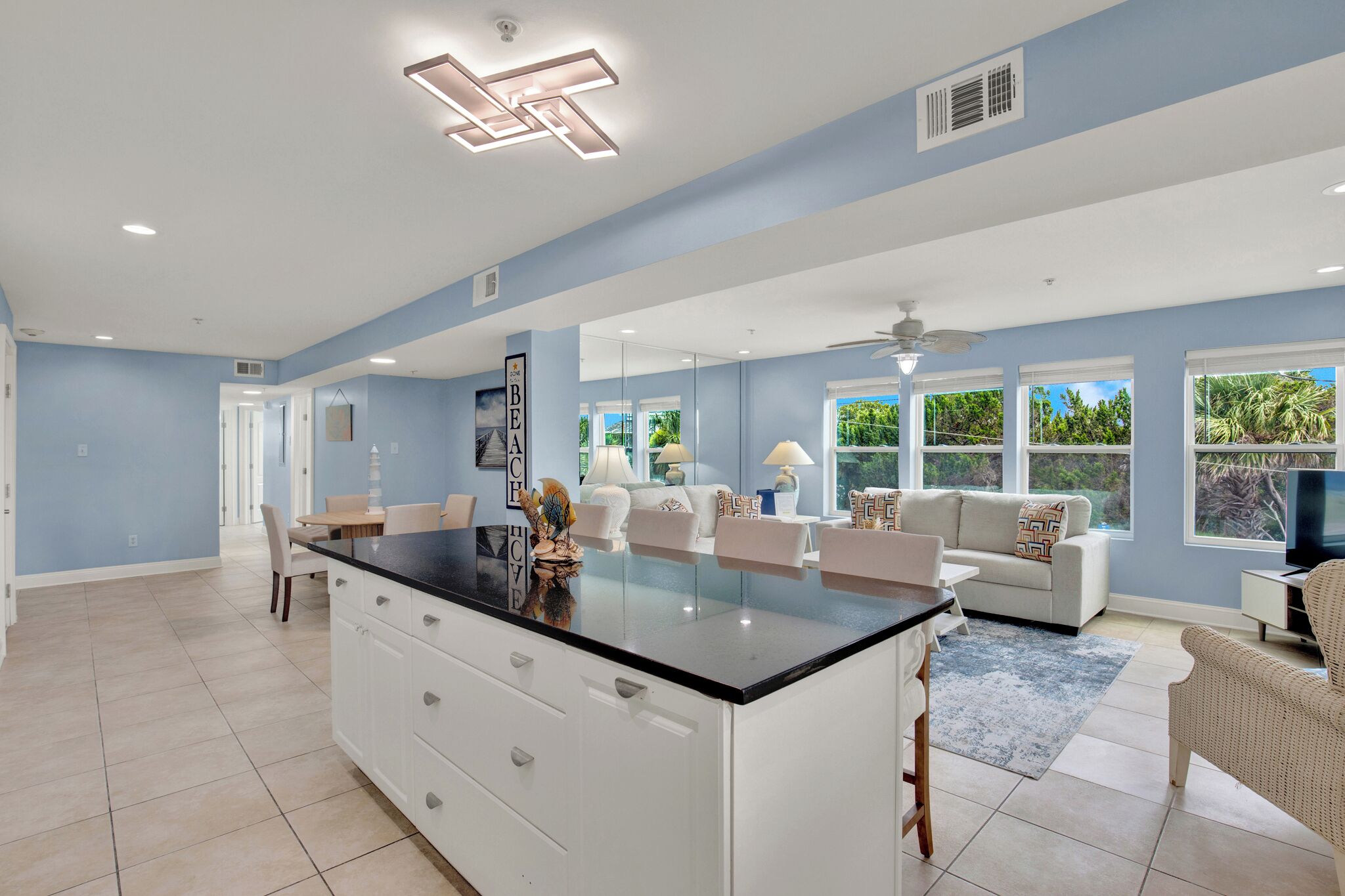 A spacious living area with a large island, white furniture, and a view of greenery outside.