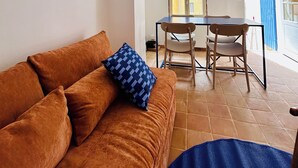 A living room with a brown sofa, a blue patterned pillow, and a blue mat. There is a dining table with chairs and a window with a view of the outside. - Charming house in the center of Aix, quiet, absolute, bright. (Aix-en-Provence)