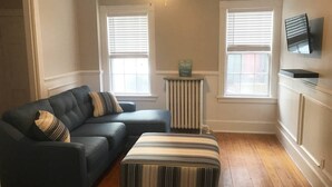 A living room with a sectional sofa, an ottoman, a ceiling fan, and a radiator.