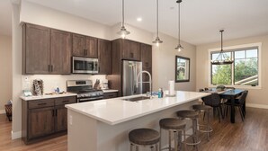 Una cocina moderna con armarios de madera oscura, electrodomésticos de acero inoxidable, una isla grande con taburetes, iluminación colgante y vistas a zonas verdes por una ventana.