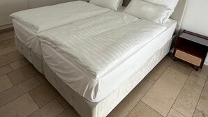 Een groot, net opgemaakt bed met witte lakens in een kamer met effen witte wanden en een betegelde vloer.