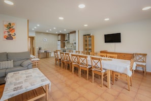 Un salon comprenant un canapé, une table basse, un coin repas avec des chaises et une table en bois, une cuisine à l’arrière-plan et un téléviseur fixé au mur.