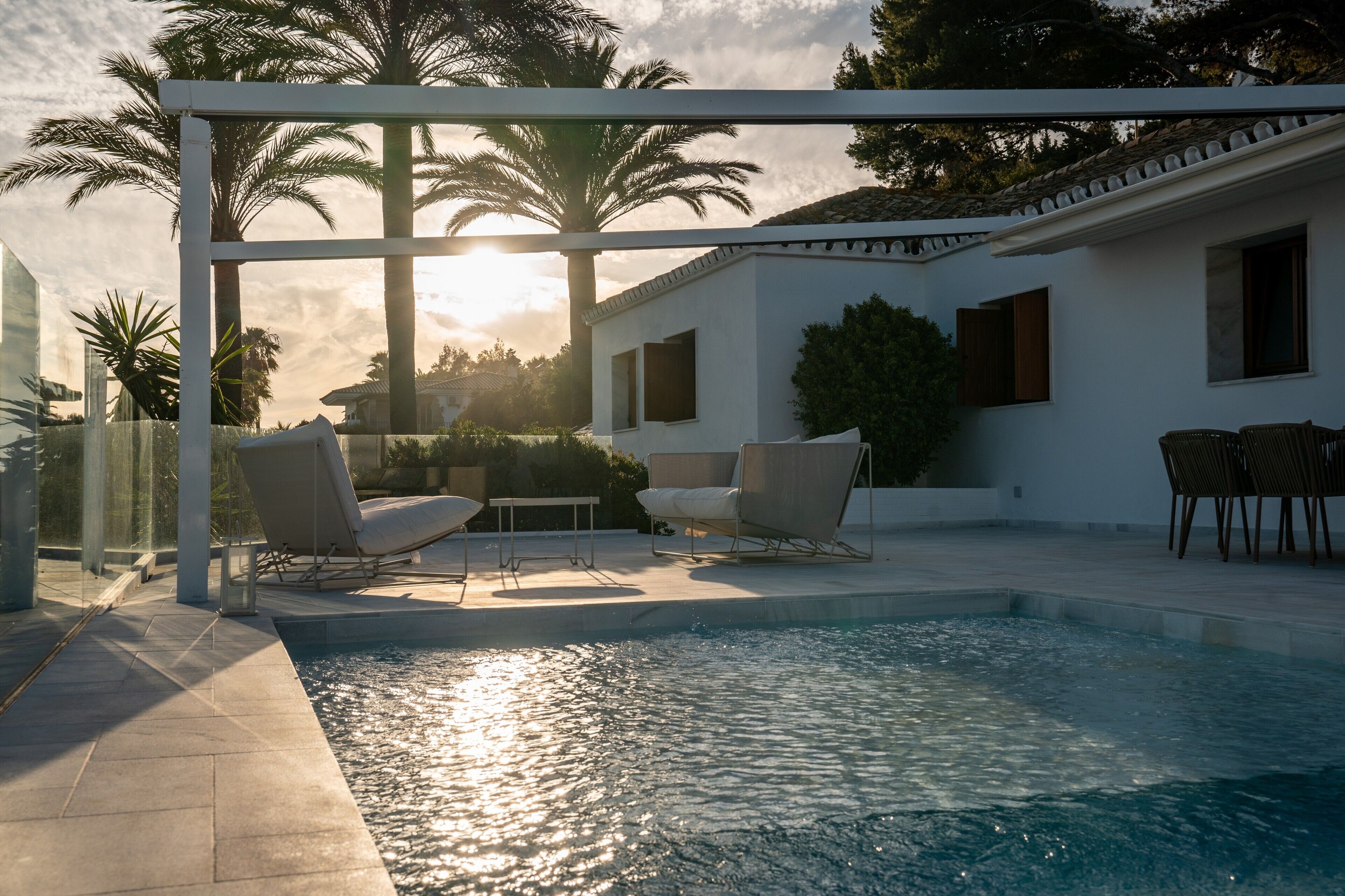 Et poolområde med liggestole, en pergola og et hus i baggrunden.