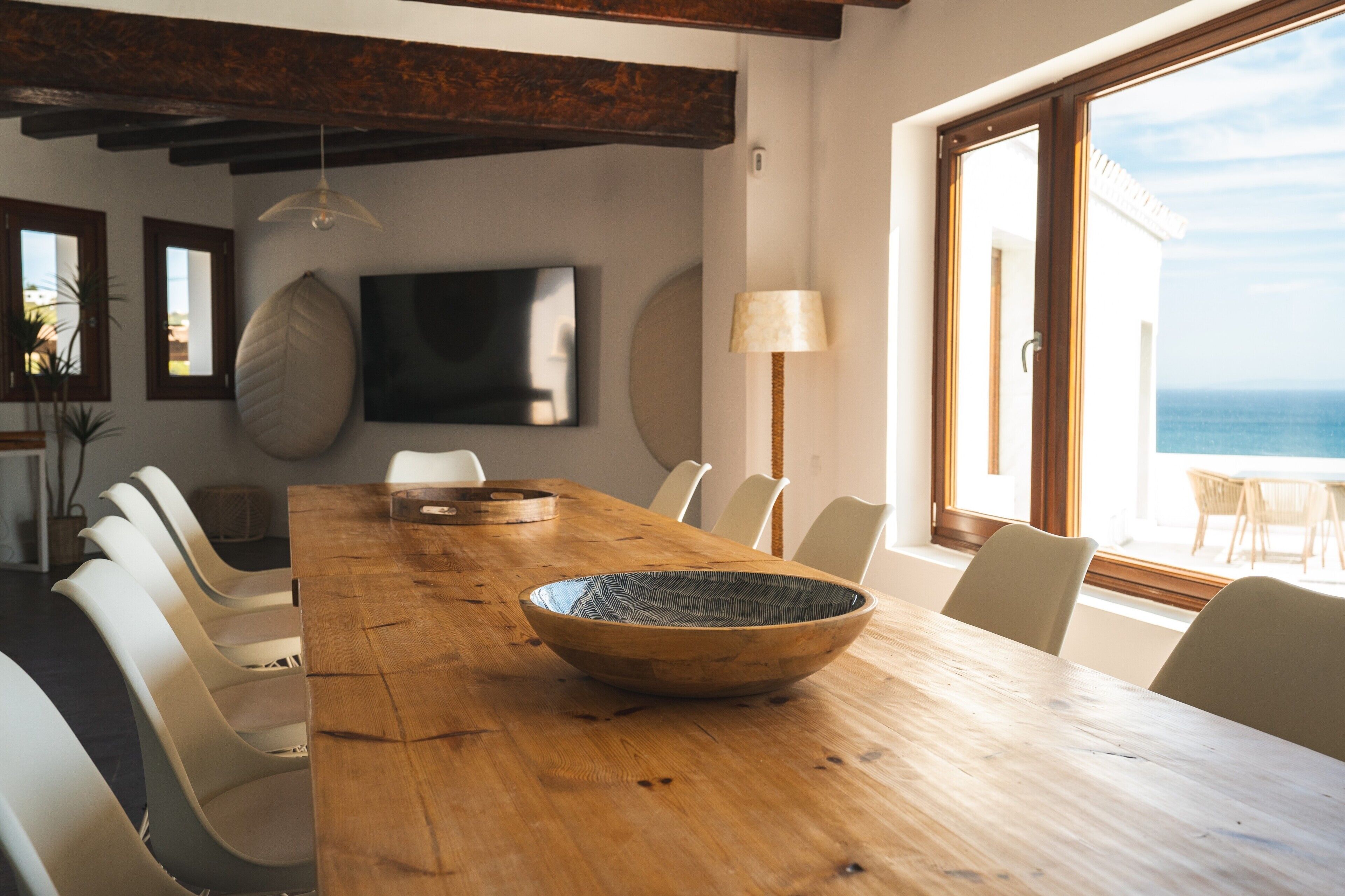 Zona pranzo con tavolo in legno, sedie, una grande ciotola intrecciata, televisore e vista sull'oceano.
