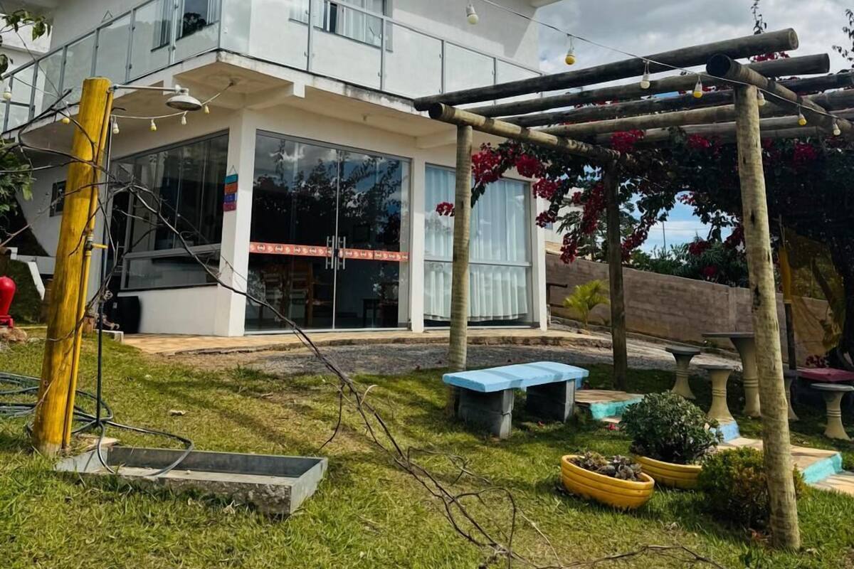 Une maison moderne à deux étages, dotée d’un balcon, d’une pergola ornée de fleurs rouges et d’un petit jardin avec des jardinières et un banc.