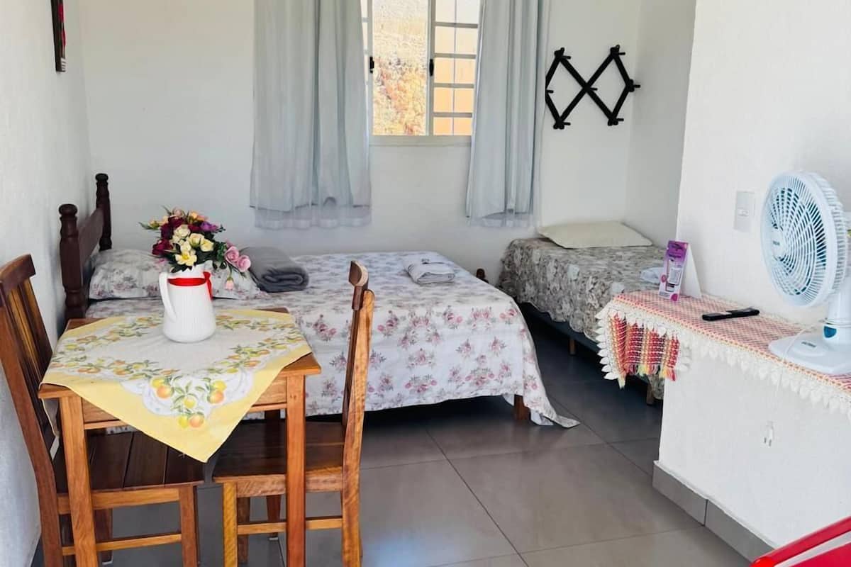 Une petite pièce propre, avec un lit, une table et des chaises, une fenêtre avec des rideaux, un ventilateur fixé au mur et une chaise rouge.
