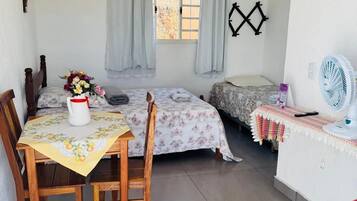 Una habitación pequeña y limpia con una cama, una mesa con sillas, una ventana con cortinas, un ventilador de pared y una silla roja.