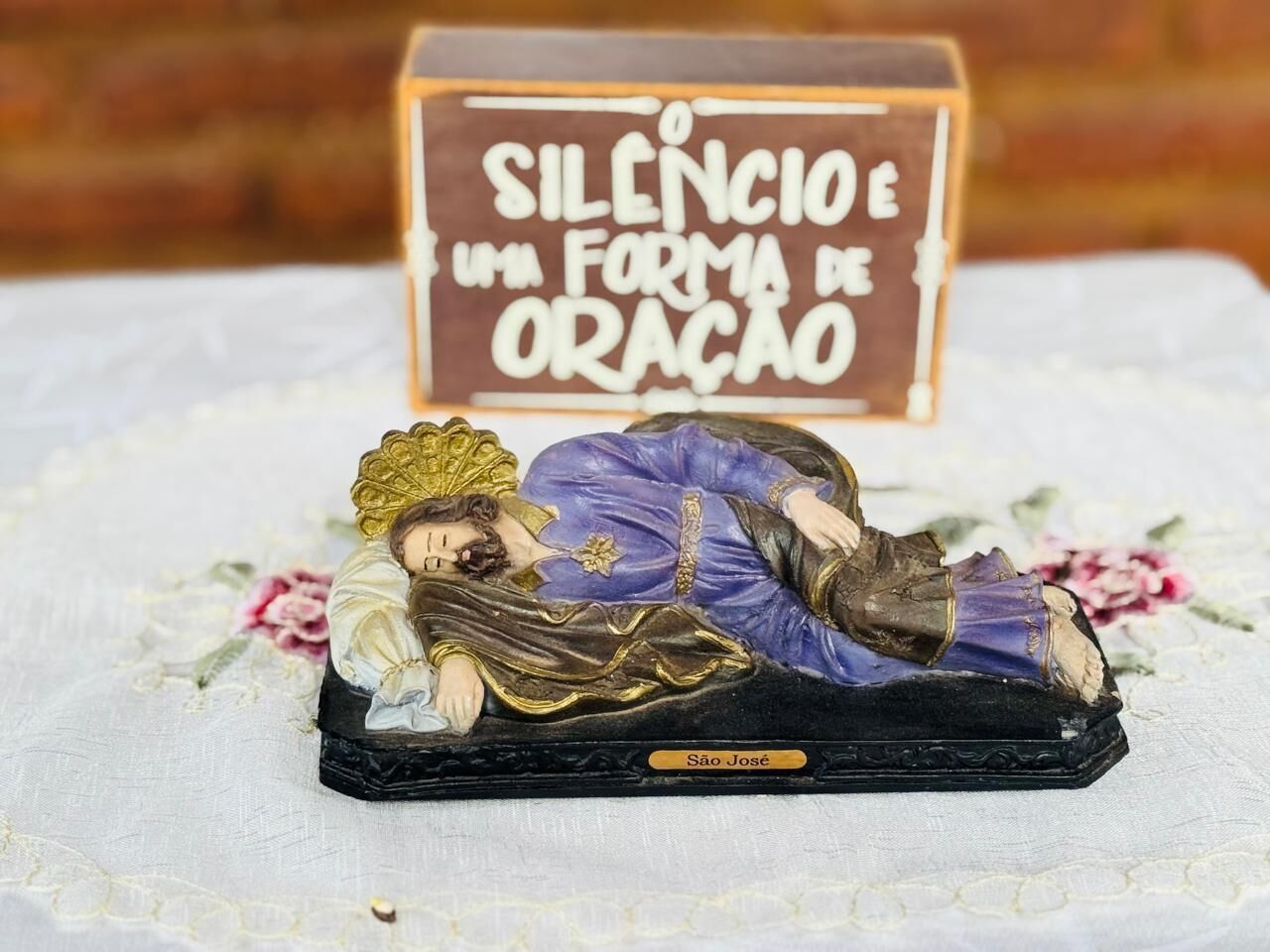 A religious figurine of São José on a table with a sign reading "SILENCIÊ UM FORMA DE ORAÇÃO" in the background.