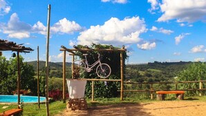 A rustic outdoor setting with a bicycle hanging on a wooden structure, a wooden bench, and a view of a lush landscape. - Pousada Dona Terezinha (Formiga)