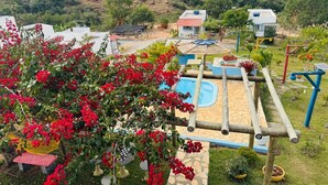 A playground with a blue slide and climbing structures, surrounded by a garden with red flowers and a view of rolling hills in the background. - Pousada Dona Terezinha (Formiga)