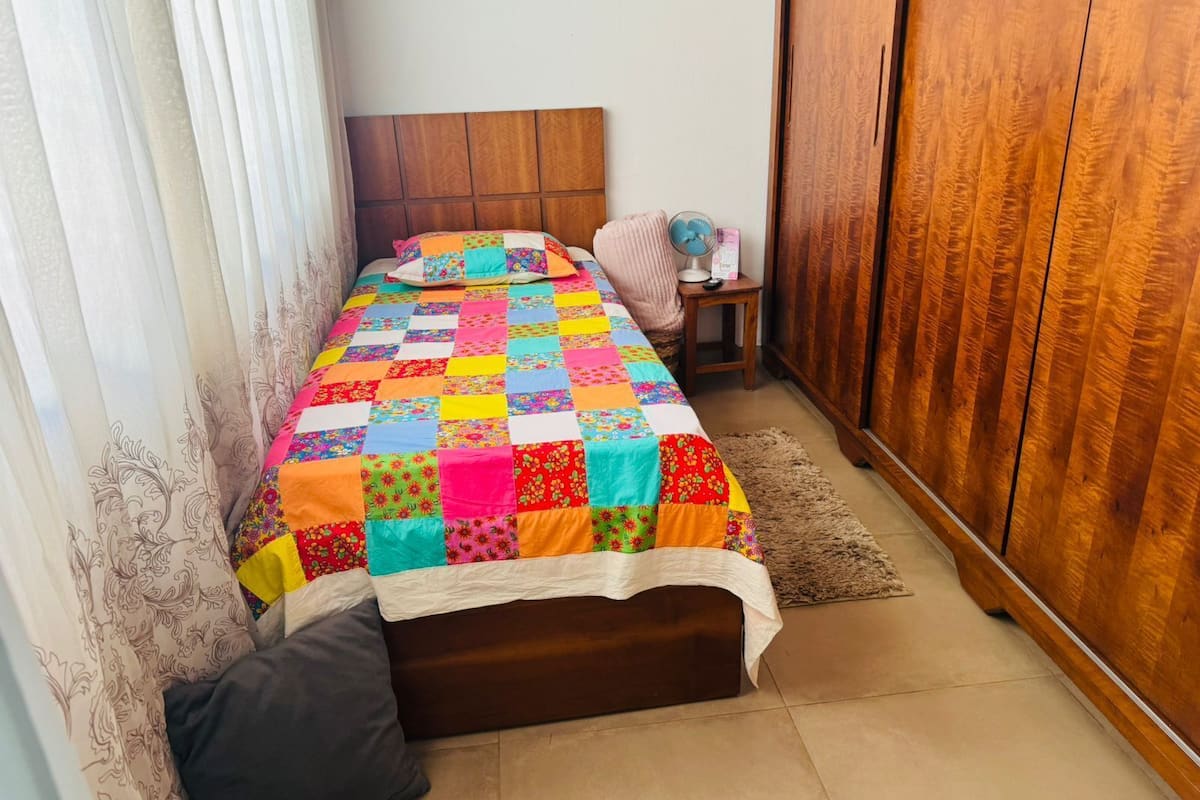 Une chambre à coucher avec un lit, une armoire en bois, une table de chevet avec un ventilateur et une fenêtre avec des rideaux.