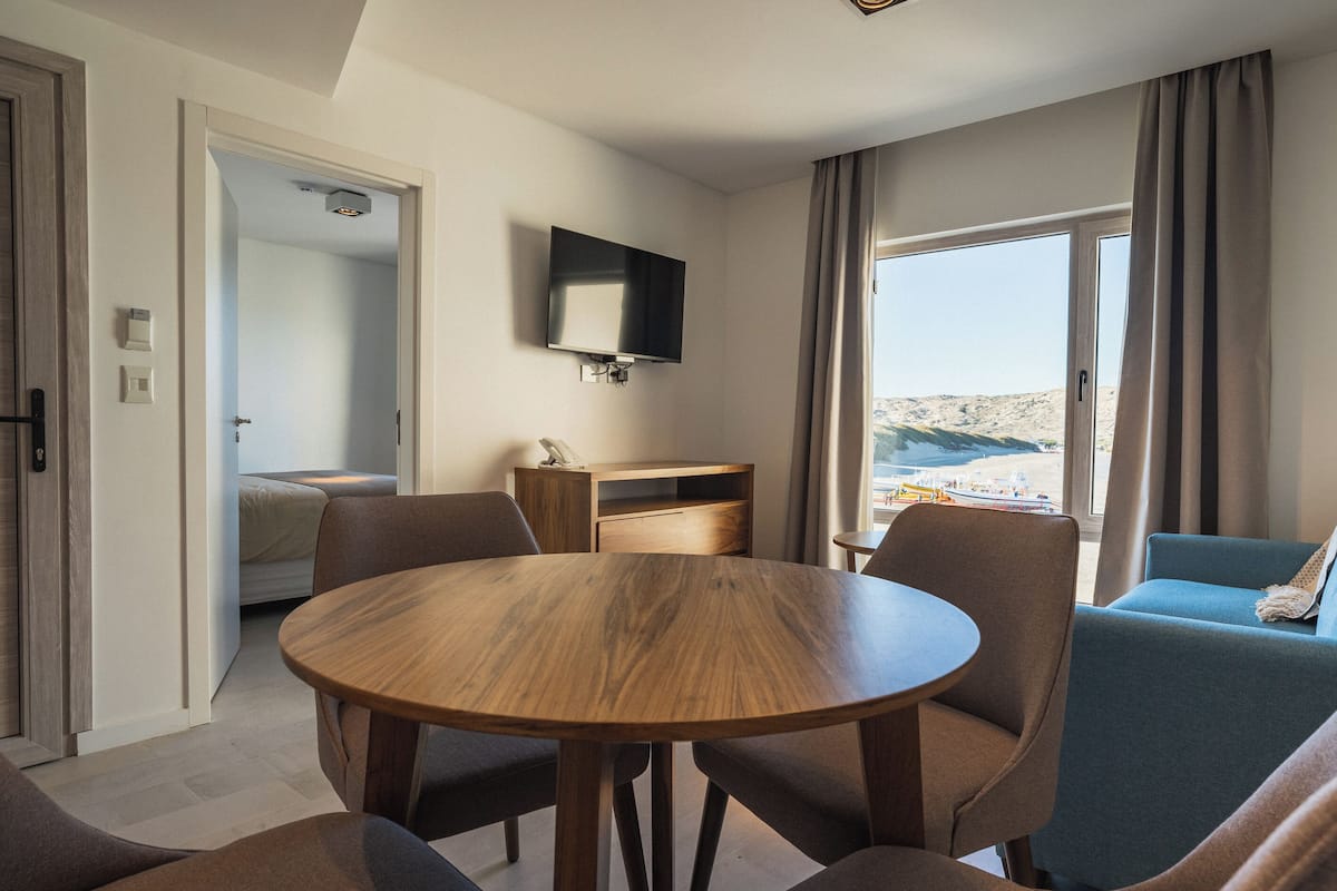 A hotel room with a wooden dining table, comfortable seating, a flat-screen TV, a small desk, and a large window offering a view of the beach.