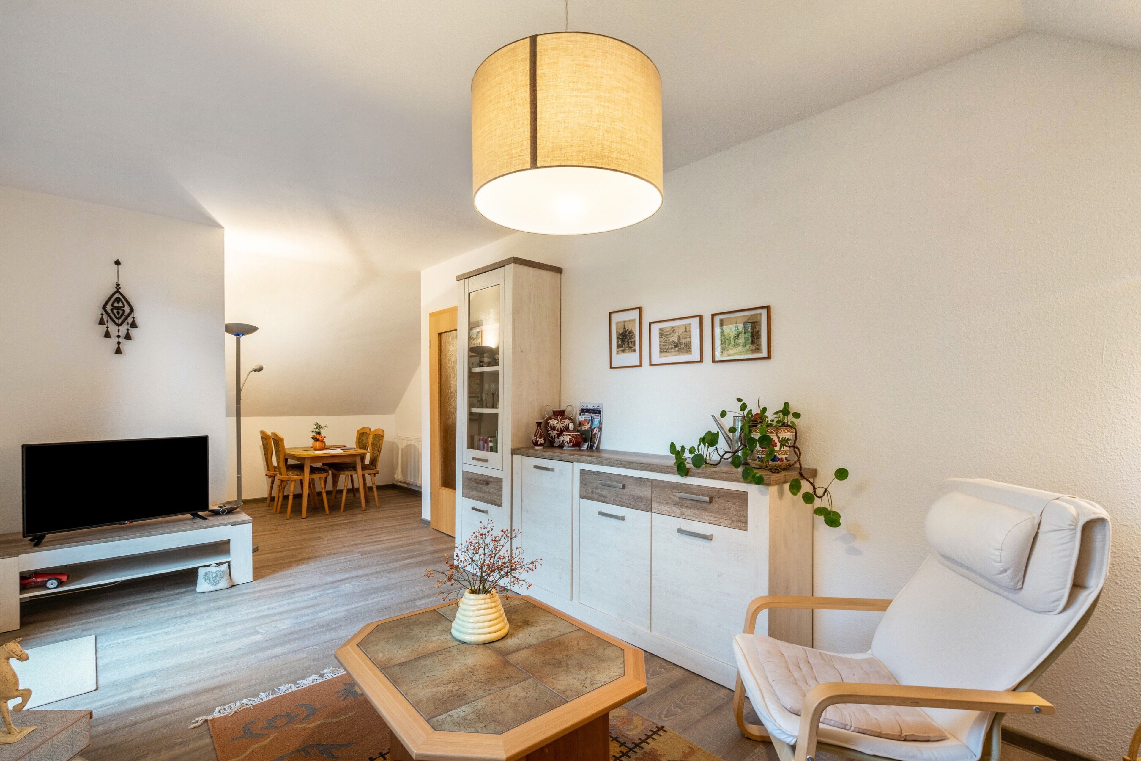A modern living room with a flat-screen TV, a dining area, a white sofa, and a hanging light fixture.