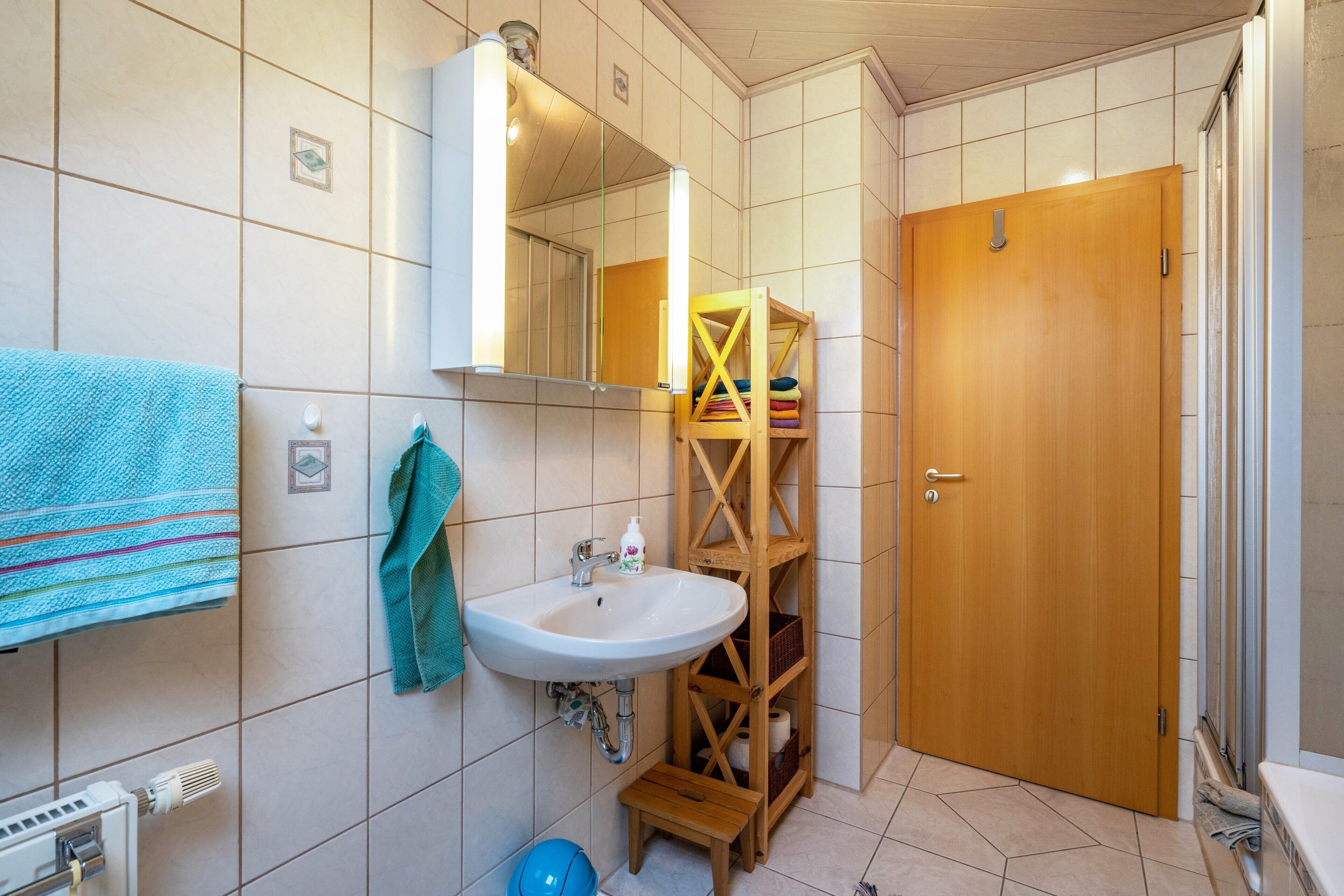 Une salle de bain comprenant un lavabo, un miroir, un porte-serviettes, une étagère en bois et une porte.