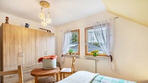 Une chambre à coucher avec un lit, une armoire en bois, une table ronde avec une chaise, et deux fenêtres avec des rideaux.