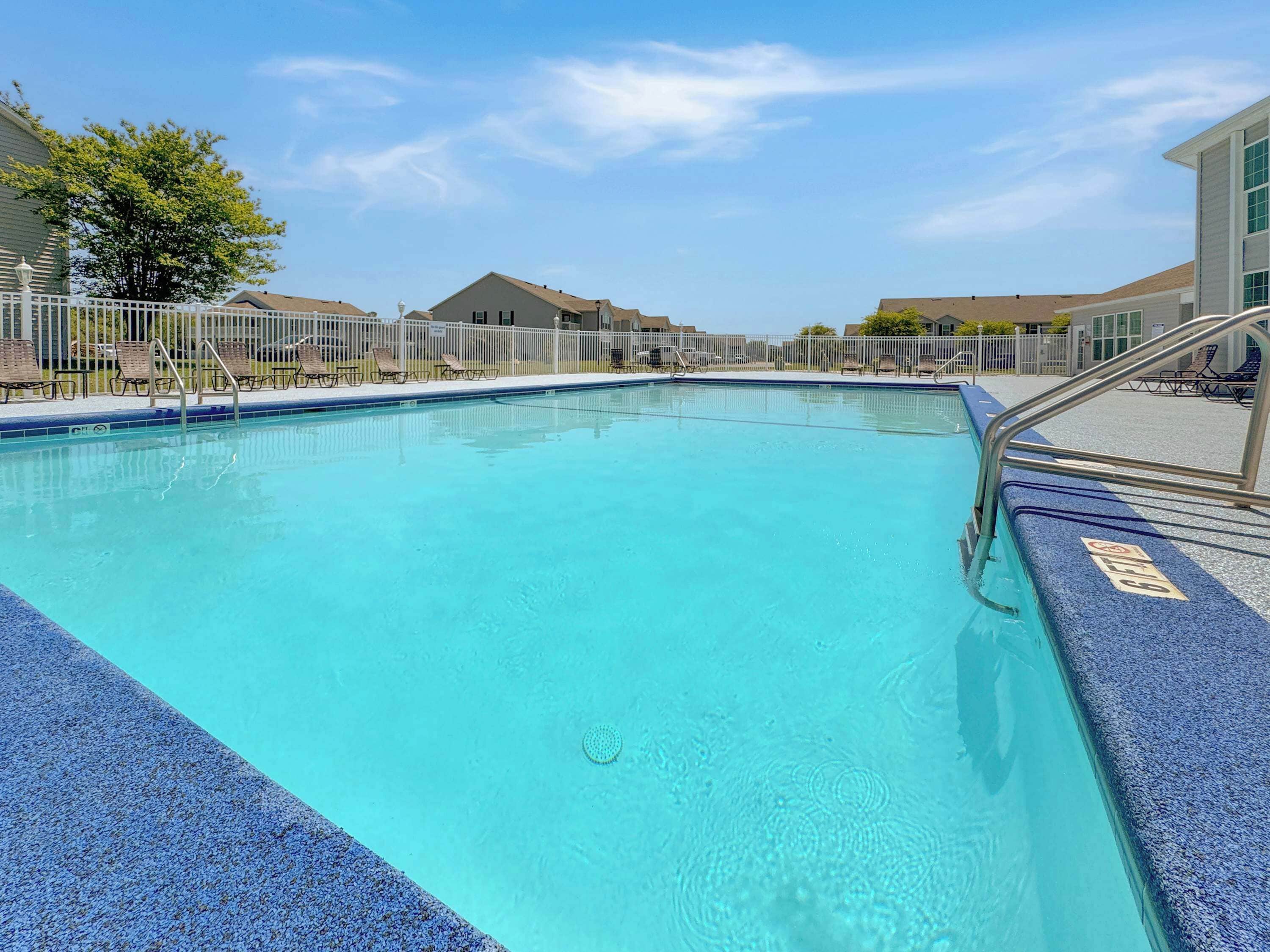 A clear blue swimming pool with a blue textured edge, surrounded by a white fence and lounge chairs.