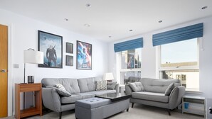 A modern living room with grey sofas, a coffee table, and a side table. There are framed posters on the wall, a window with blue curtains, and a door. - Pass the Keys |Modern 2bed Apartment Edinburgh (Edinburgh)