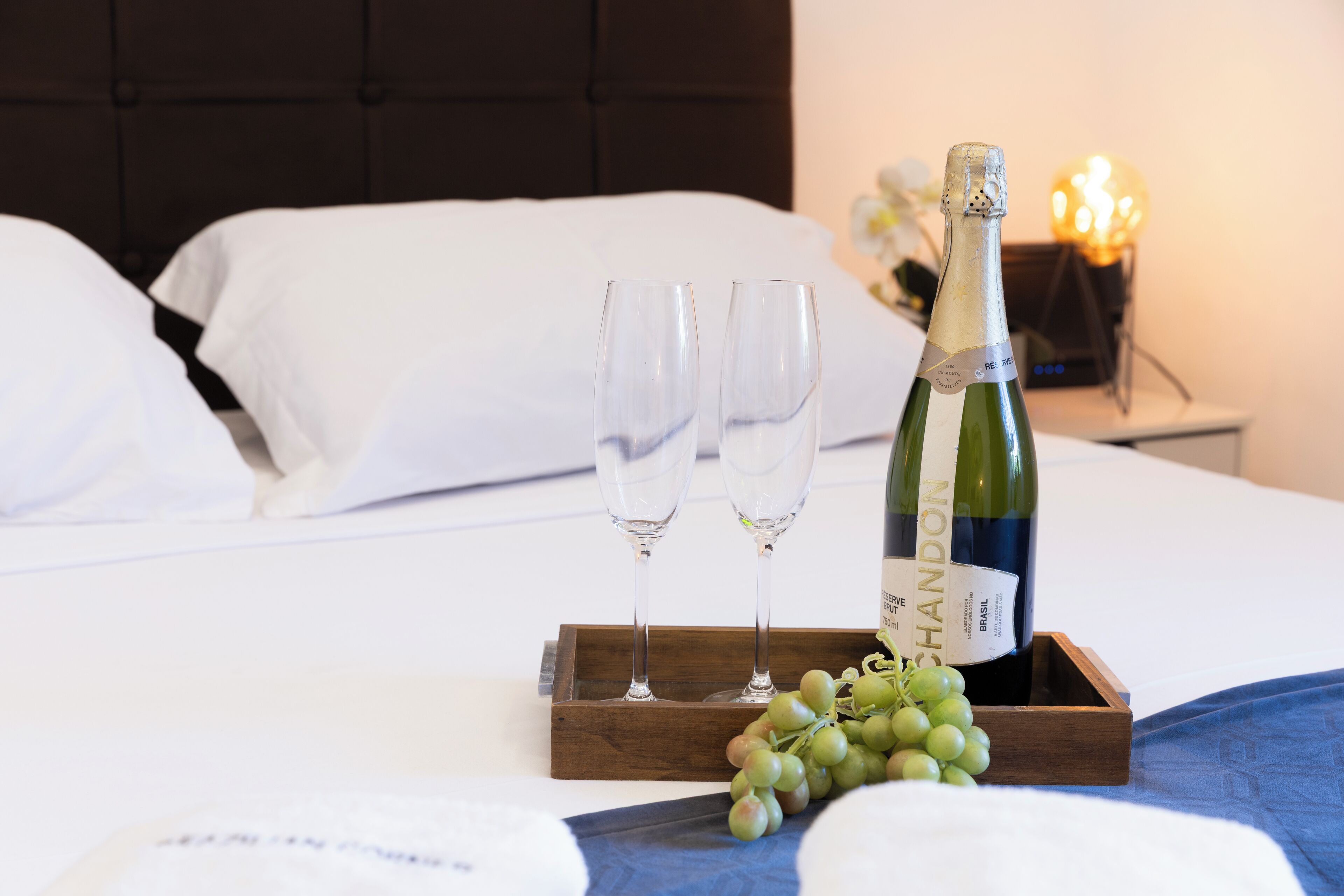 A bed with white linens, a wooden tray with a bottle of Chandon champagne and two glasses, and a bunch of grapes.