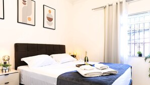 A neatly made bed with a dark headboard, white linens, and a folded towel on the side. There is a bottle of champagne and two glasses on the bed.