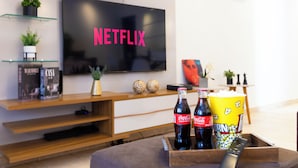 A modern living room with a flat-screen TV displaying 'NETFLIX', a wooden entertainment unit with books and decorative items, and a popcorn bucket on a tray.