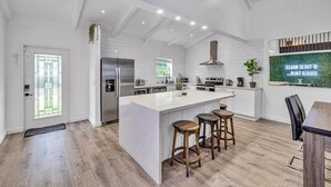 Une cuisine moderne avec des meubles blancs, un grand îlot central doté de tabourets, des appareils électroménagers en acier inoxydable et une vue sur l’extérieur grâce à une fenêtre.