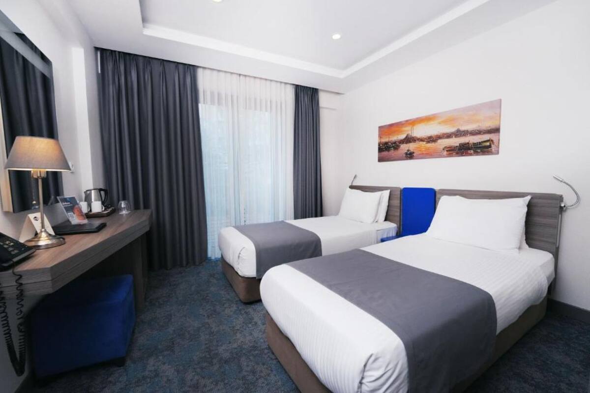 Habitación de hotel con dos camas, un escritorio con lámpara, cafetera y un cuadro en la pared.