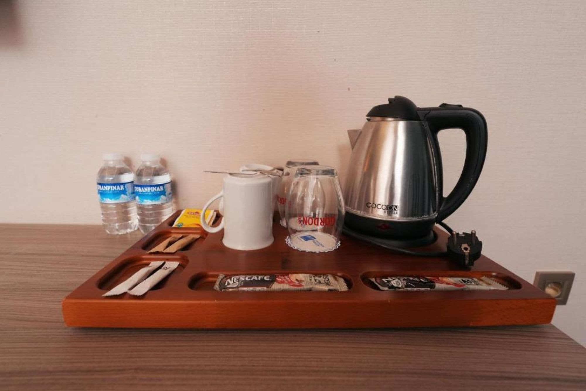 A wooden tray with a coffee maker, water bottles, a glass, a mug, and some snacks.
