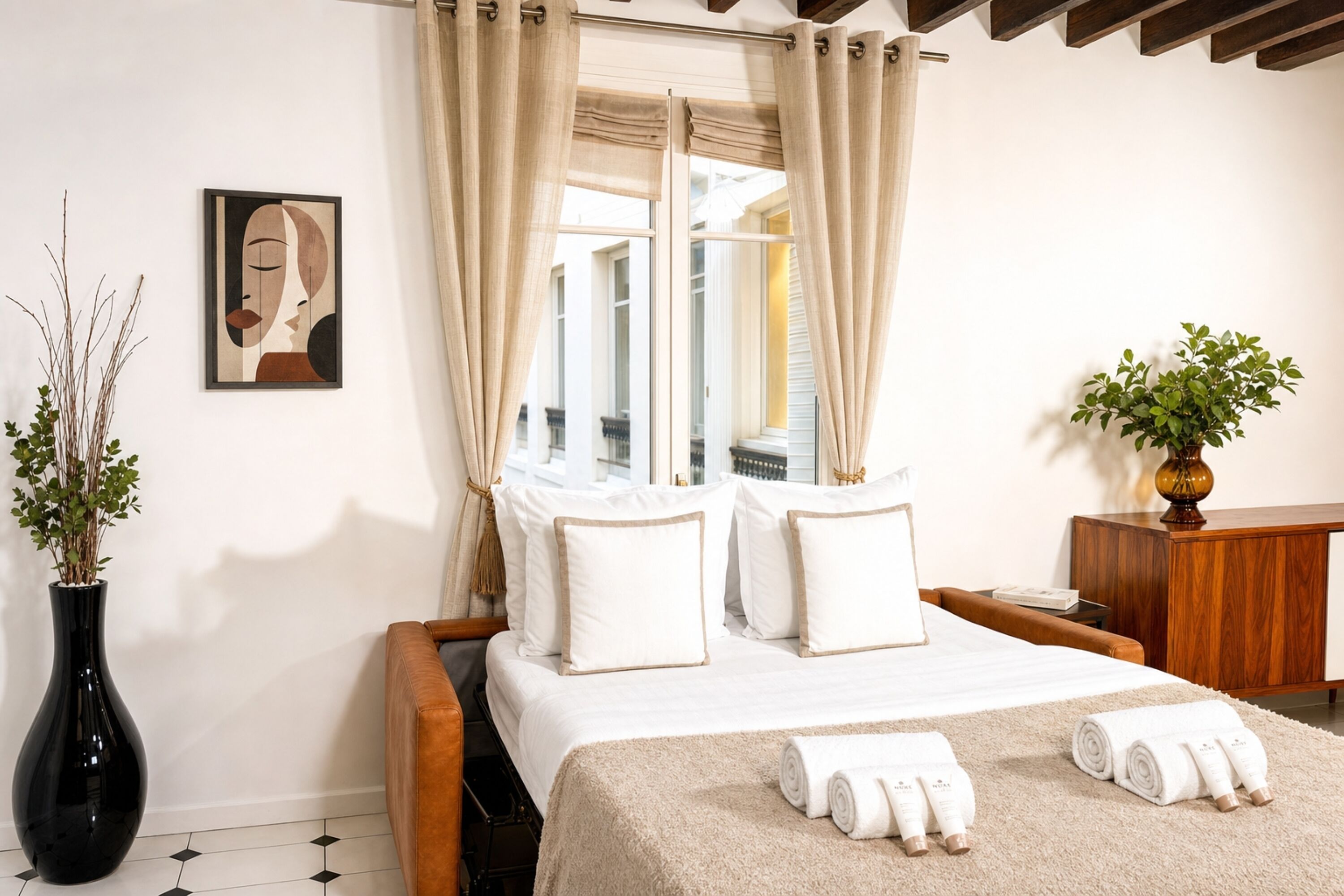 A hotel room with a bed, curtains, a vase with plants, a painting, and a wooden cabinet.