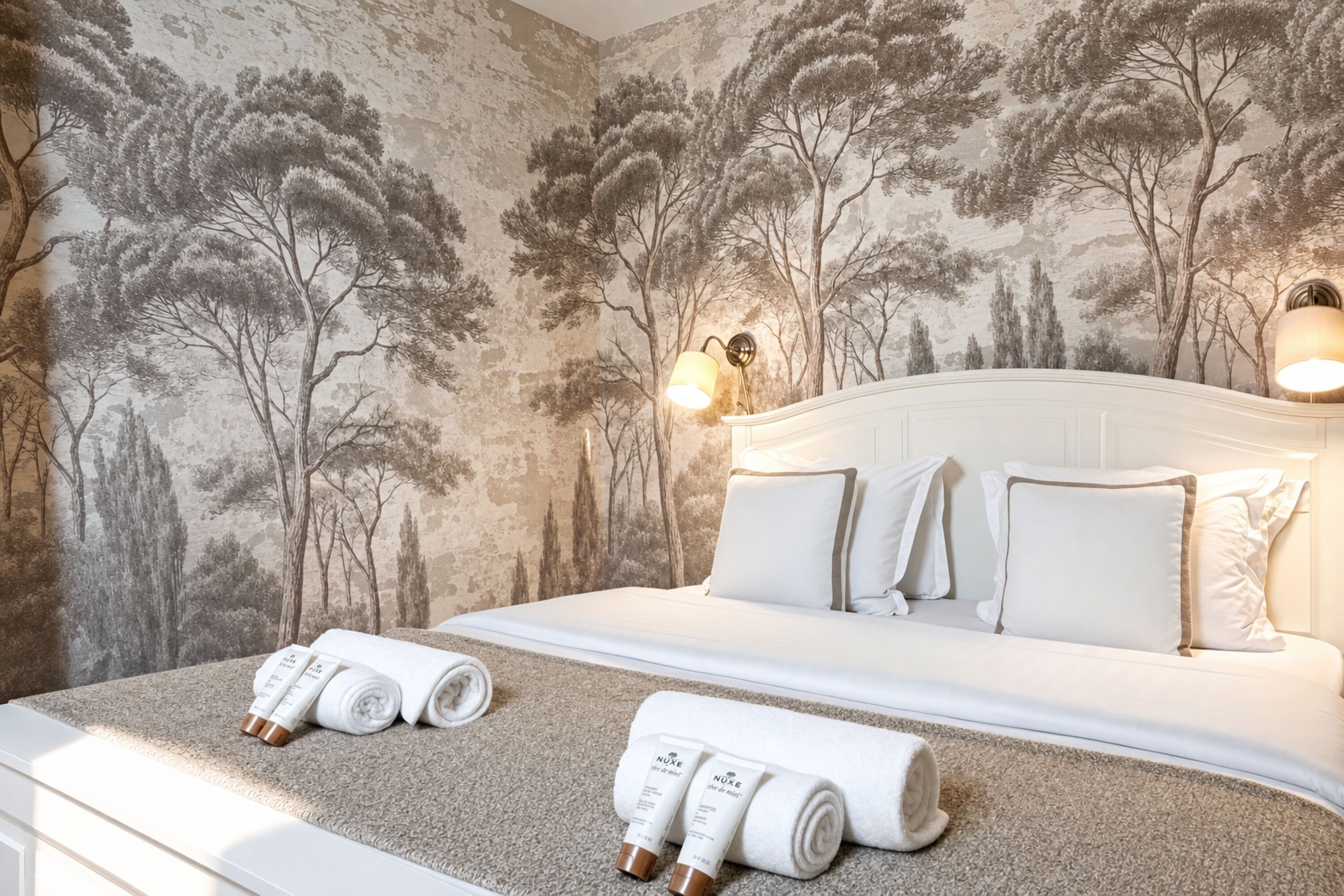 Un lit bien fait, avec du linge de lit blanc et des oreillers, une tête de lit agrémentée de lampes murales et un mur décoré d’une fresque représentant un arbre.