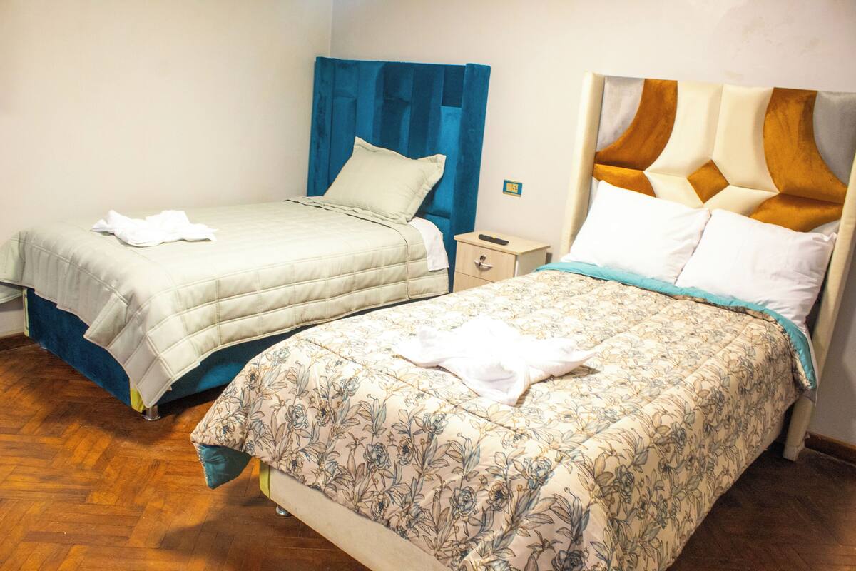 Two beds with different headboard designs, a wooden floor, and a small bedside table with a remote control.