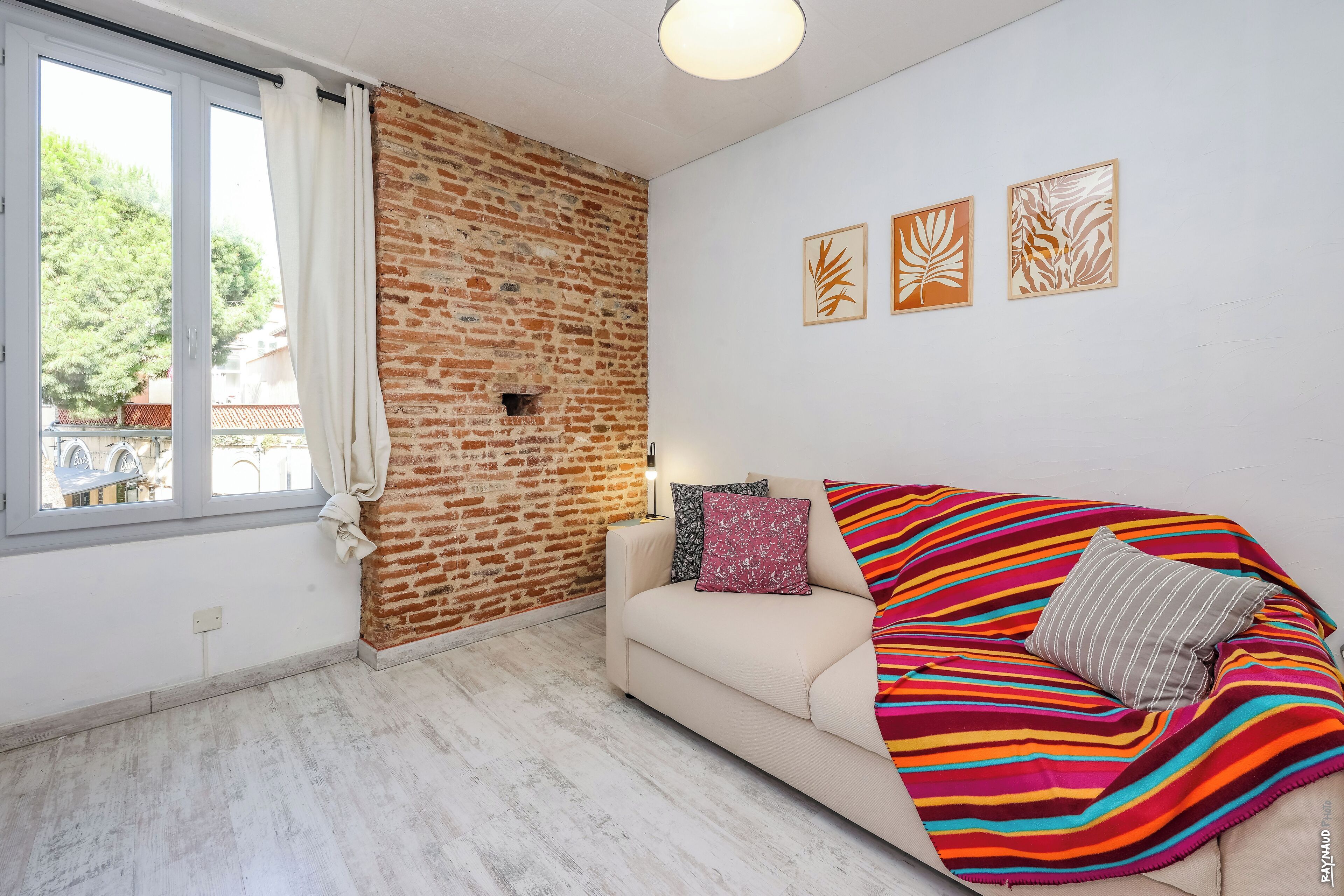 Una sala de estar moderna con una pared de ladrillo decorativa, un sofá con cojines coloridos y tres obras de arte con motivos de hojas.