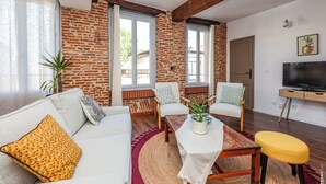 A living room with a brick wall, white sofa, and a TV on a wooden stand. - Gambetta Apartment – Right in the Heart of Gaillac (Gaillac)