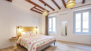 A bedroom with a wooden floor, a bed with floral bedding, a bedside table, a hanging light fixture, and a window with curtains. - Gambetta Apartment – Right in the Heart of Gaillac (Gaillac)