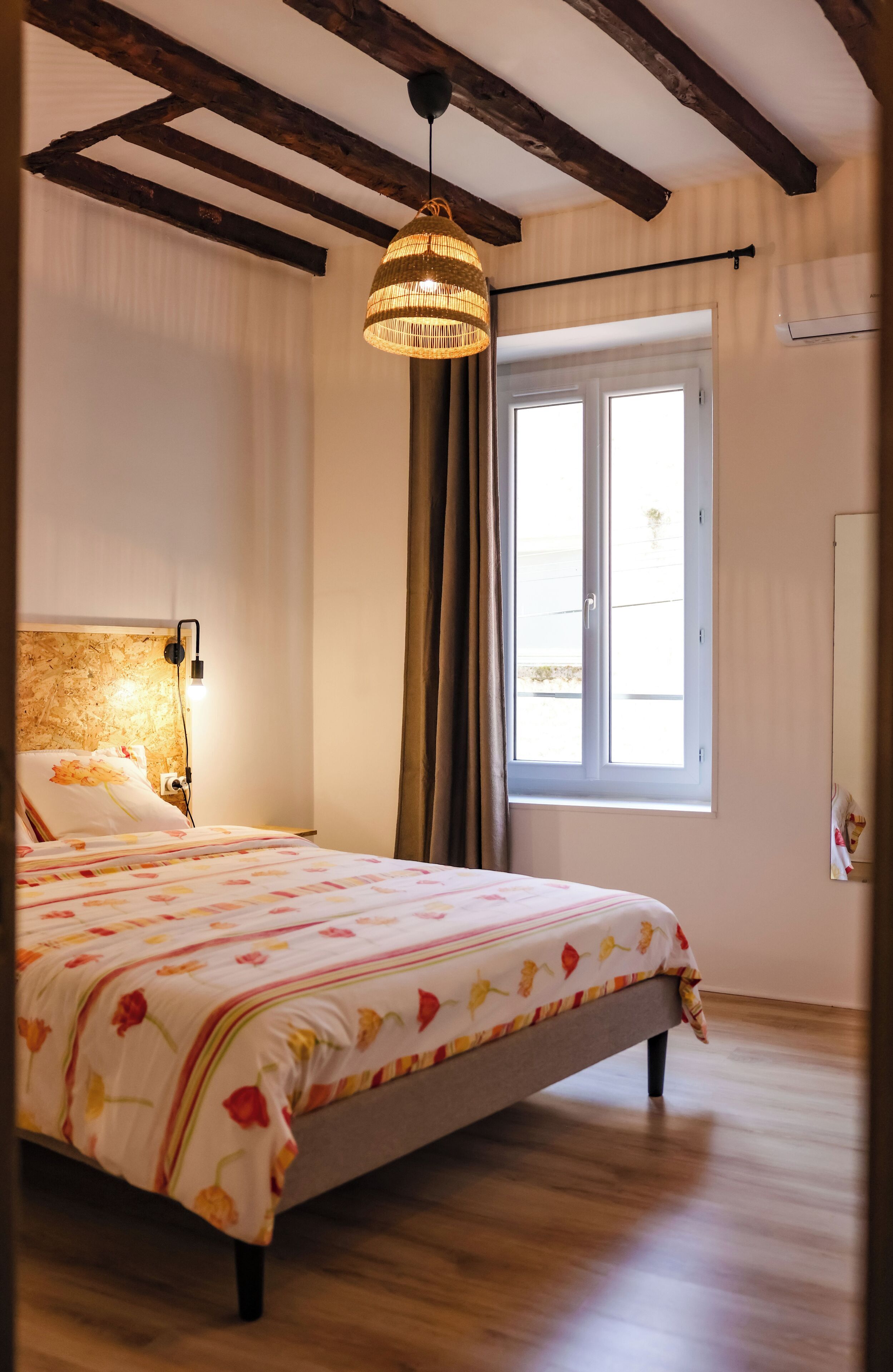 Una camera da letto con un letto, una lampada a sospensione, una finestra con tende e travi in legno al soffitto.