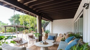 Een overdekt terras met rieten meubels, een ronde tafel en diverse planten.