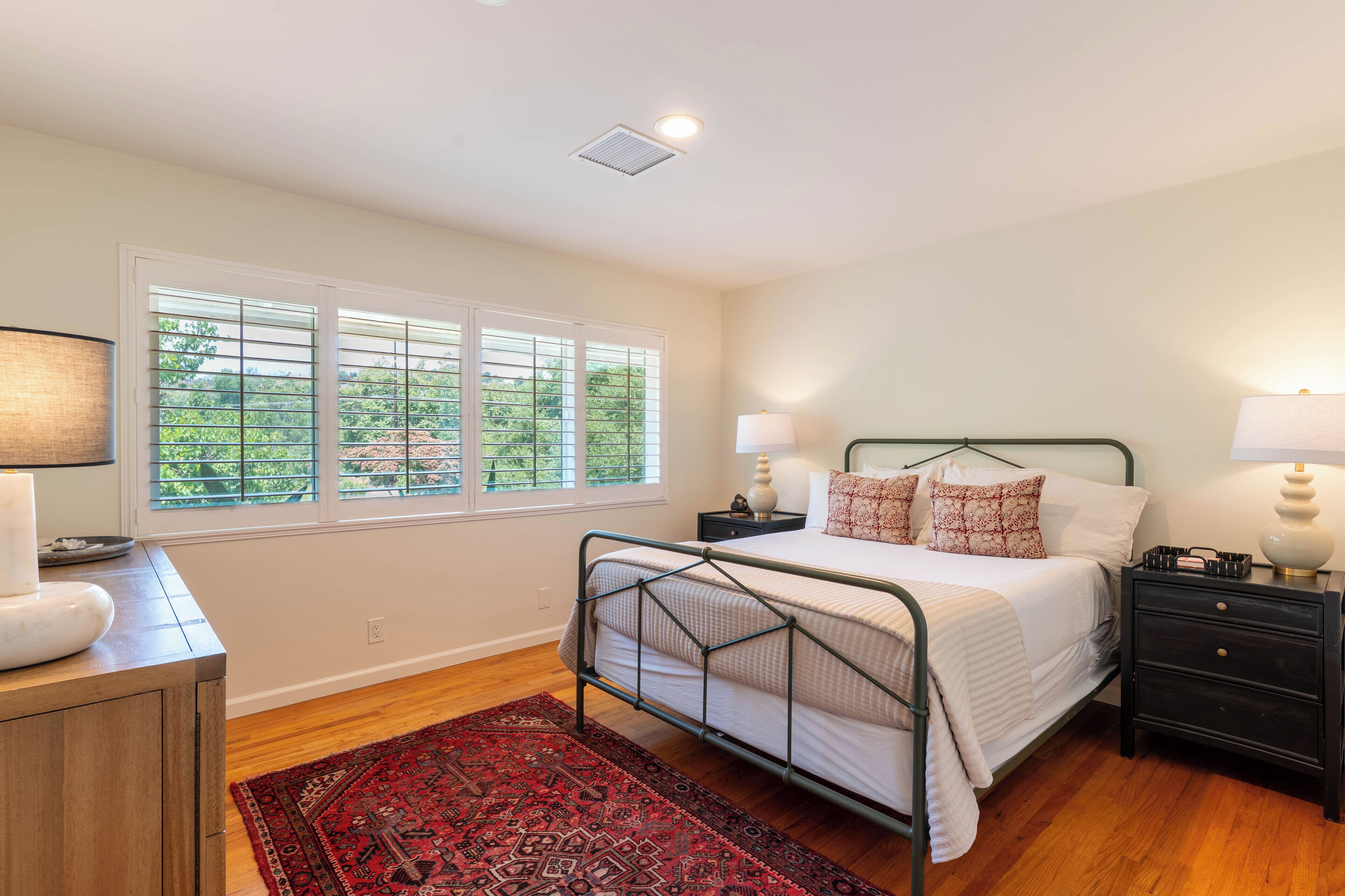 A bedroom with a bed, bedside tables, a nightstand, a dresser, a window with blinds, and a red patterned rug.