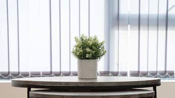 A round table with a black metal frame and a white pot with green plants on top, placed on a wooden floor in front of a window with vertical blinds.