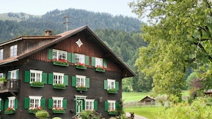 En traditionell chalet med många fönster och balkonger, omgiven av frodig grönska och med ett bergigt landskap i bakgrunden.