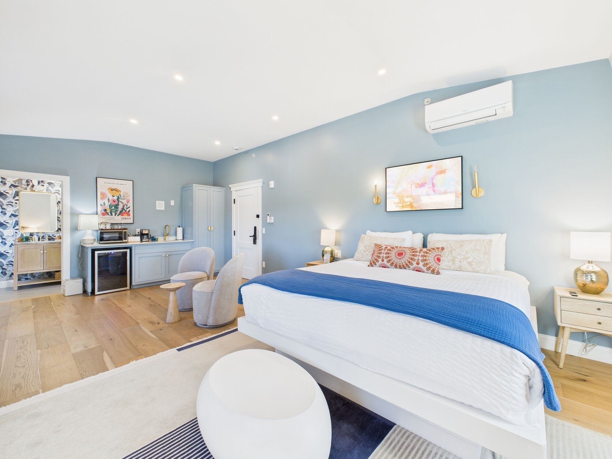 A bedroom with a bed, bedside tables, a desk, chairs, and an air conditioning unit.