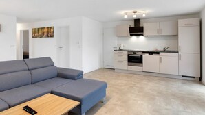 A modern living room with a grey sofa and a wooden coffee table, a kitchen with white cabinets and a black range hood, and a painting on the wall.