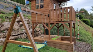A wooden playhouse with a swing set and a slide, surrounded by a landscaped garden.