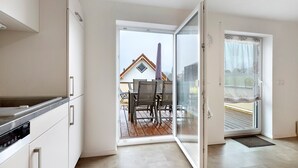 An open sliding door leading to a balcony with a dining table and chairs, a kitchen area with cabinets and a stove, and a view of a house with a sloped roof.