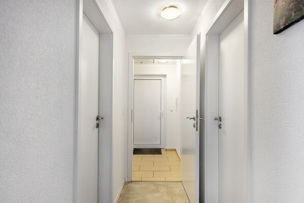 A narrow hallway with white walls and doors, a tiled floor, and a recessed ceiling light.