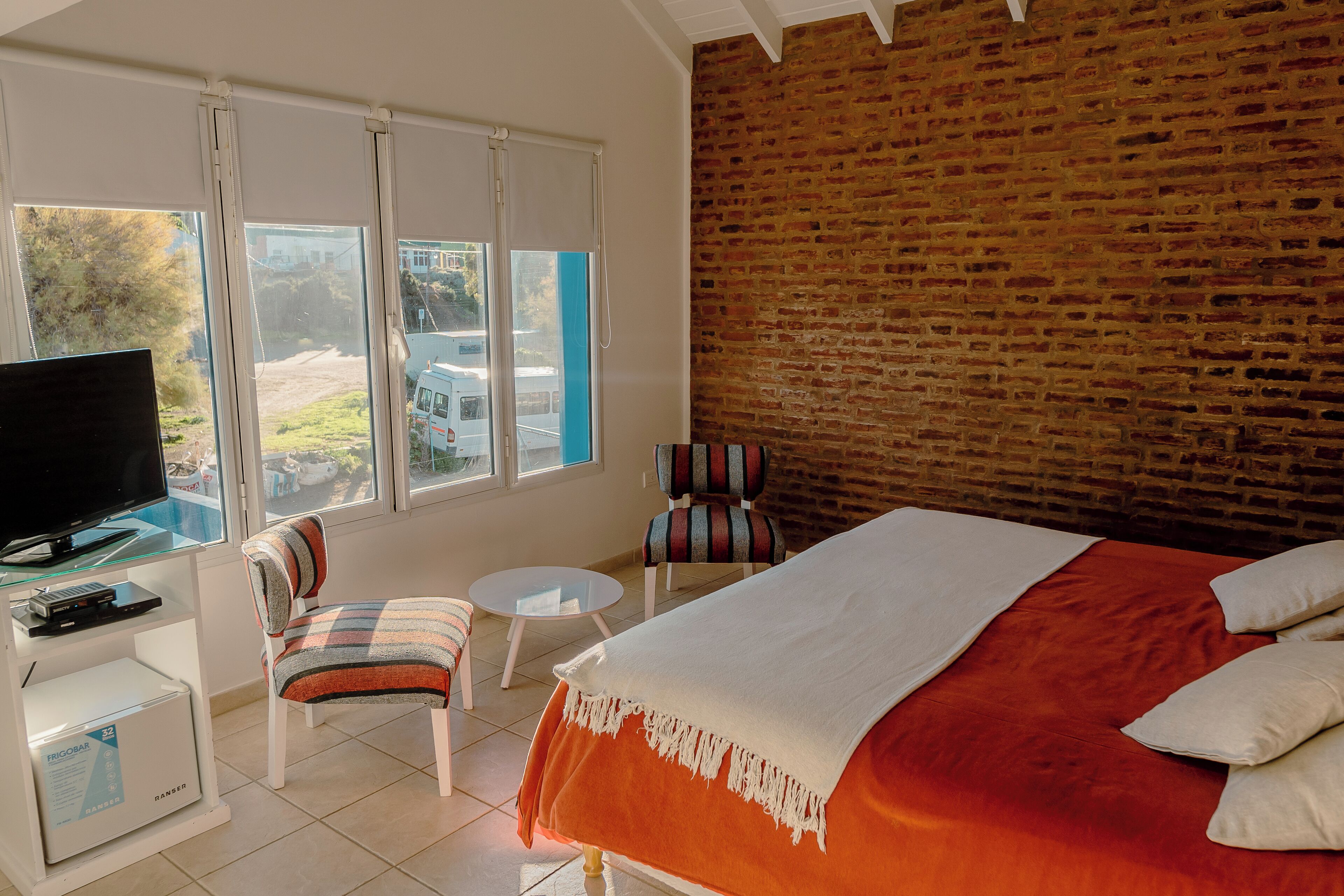 Une chambre d’hôtel avec un mur en briques, un lit recouvert d’une couette rouge et blanche, un téléviseur posé sur un meuble blanc, un fauteuil à rayures et une petite table ronde.