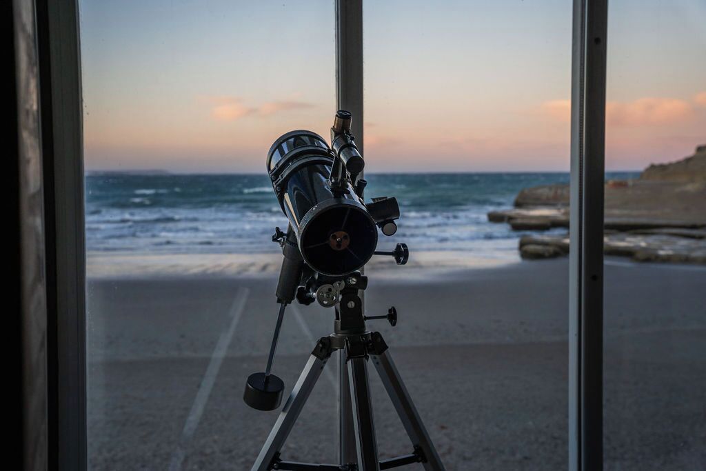 Un télescope posé sur un trépied, près d’une fenêtre donnant sur une plage.