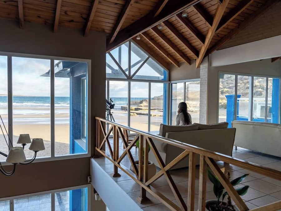 Une pièce spacieuse avec de grandes fenêtres donnant sur une plage, des poutres en bois au plafond et un balcon doté d’une balustrade.