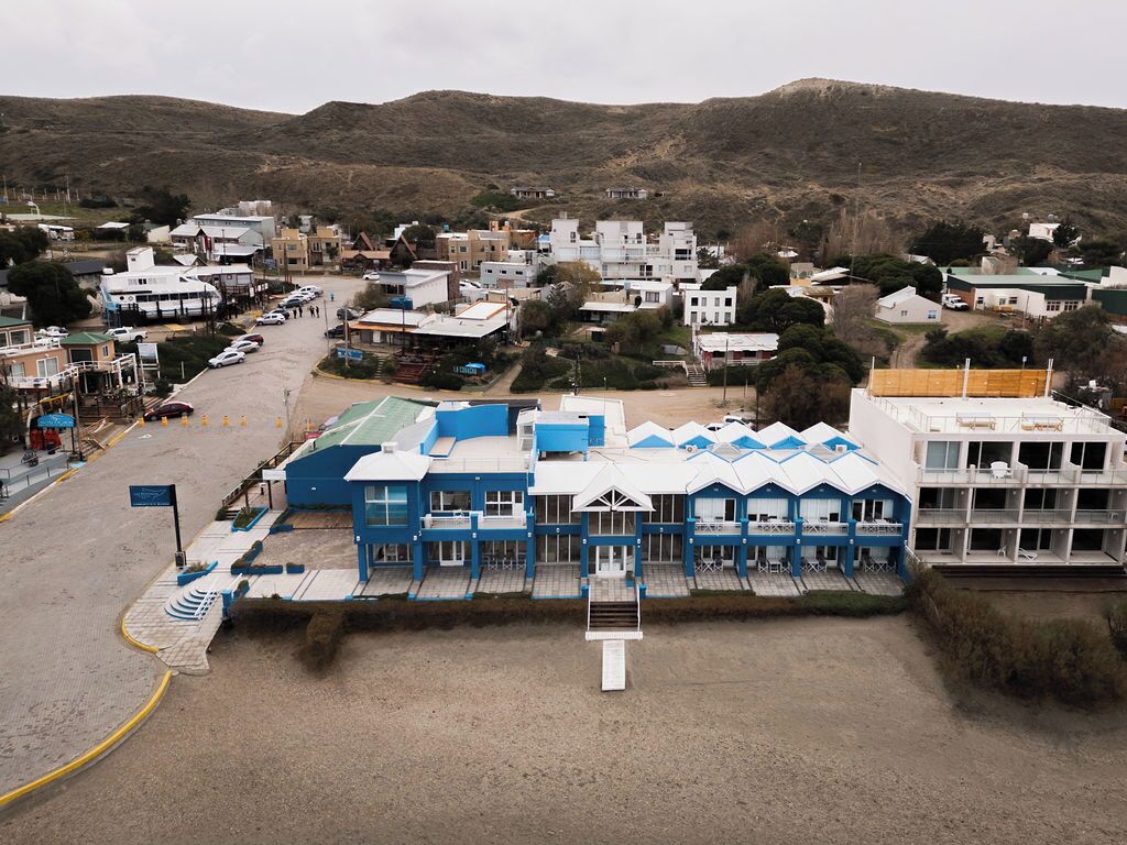 A coastal hotel with multiple blue and white buildings, a parking area, and a view of the surrounding hills.