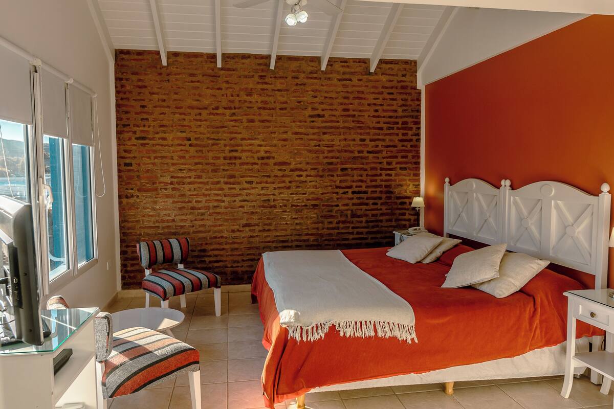 Ein Schlafzimmer mit Backsteinwand, einem Bett mit weißem Kopfteil, einem gestreiften Sessel und einem Glastisch.
