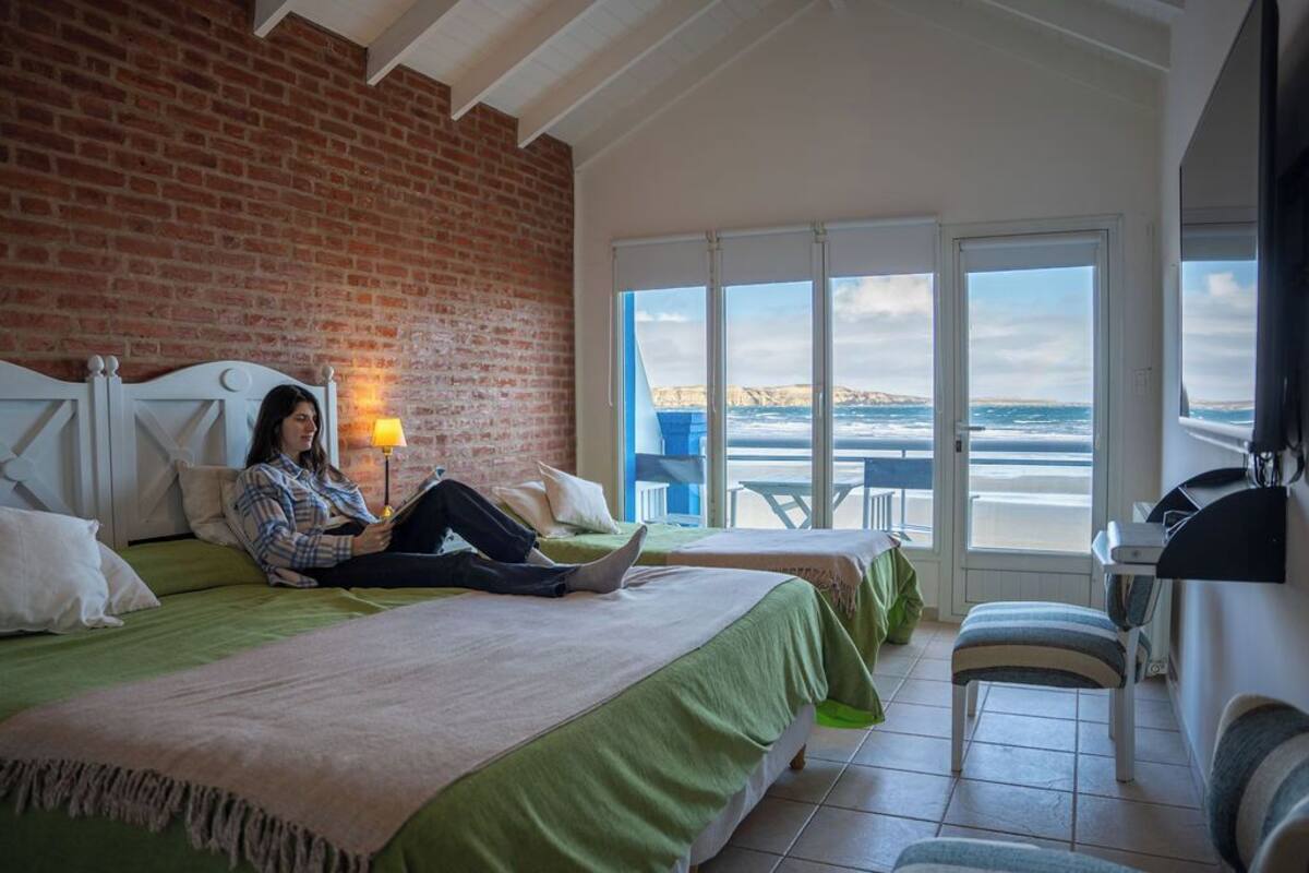 Ein Schlafzimmer mit Backsteinwand, einem großen Fenster mit Meerblick, einem Bett mit grüner Bettwäsche und einem Sessel.
